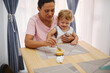 © Taras Grebinets - Happy toddler sitting with mother while learning to eat at the dining table with joy and family bonding