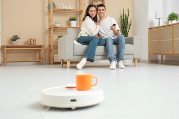 Wall Mural - Young couple using mobile phone for remote control of modern robot vacuum cleaner with cup in living room