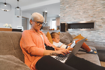 Blonde woman working from home on her laptop while her son is doing homework on a digital tablet sitting nest to her on the couch