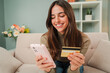 © Jose Calsina - Young caucasian woman using a smart phone to make an online purchase with a credit card sitting at home sofa. Female looking for a delivery with a cellphone at living room couch. Girl shopping online