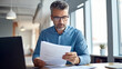 © halo - A man in a blue shirt and glasses reads a document while sitting at a desk with a laptop, in an office setting.