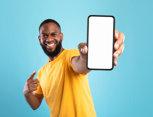 Poster - Excited black guy in casual wear pointing at cellphone with empty screen on blue studio background, space for website or mobile app design. Cellphone display mockup. Selective focus