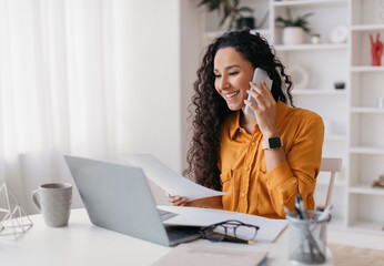 Wall Mural - Cheerful Lady Talking On Cellphone Holding Paper Sitting At Desk And Using Laptop Computer Working Online In Modern Office. Entrepreneurship And Business Career, Distance Freelance Job Concept