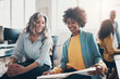 © Flamingo Images - Diverse businesswomen smiling while going over paperwork in an office. Behind them, stand two colleagues talking at a computer
