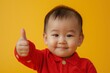 © Instacraft.Studio - A close-up portrait of a cute baby boy giving a thumbs up, wearing a red shirt against a bright yellow background. He has a big smile on his face and seems very happy.