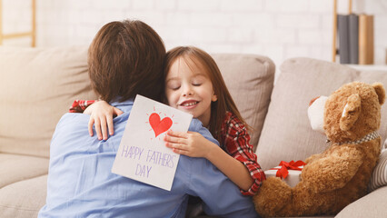 Wall Mural - Cute little preschool girl greeting her dad with Father's Day, hugging daddy with gift card in her hand, free space