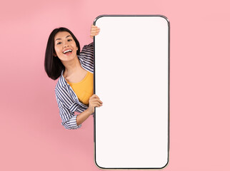 Poster - Advertising Concept. Portrait of excited asian lady peeking out big giant vertical smartphone with white blank screen presenting device display. Gadget with empty free space for mock up, pink wall