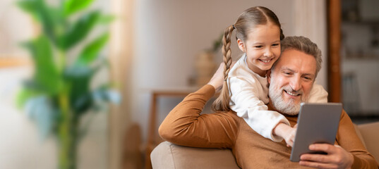 A grandfather and his granddaughter enjoy a happy moment together, laughing as they explore a tablet. The cozy living room, with soft lighting, creates a warm atmosphere.