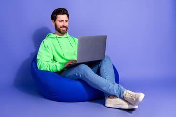 Sticker - Full length photo of handsome young guy sit beanbag hold netbook dressed stylish green garment isolated on violet color background