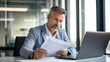© halo - Photo of a middle-aged businessman sitting at his desk in an office, looking over papers with a thoughtful expression while using a laptop computer.