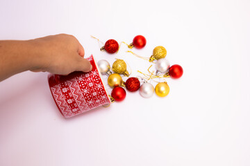 Female hand holding a red Christmas mug and scattering gold, red, and silver Christmas balls on a white background with top view