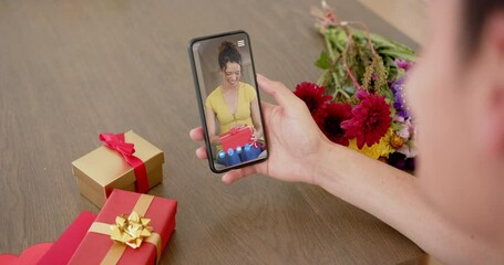 Wall Mural - Biracial woman holding smartphone with woman talking on screen with gifts on desk