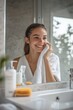 © fotofabrika - Young woman enjoying a morning skincare routine in a bright bathroom while smiling in front of a mirror