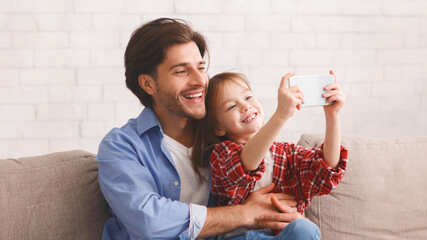 Wall Mural - Preschool girl taking selfie with her cheerful father, sitting on sofa at home, copy space