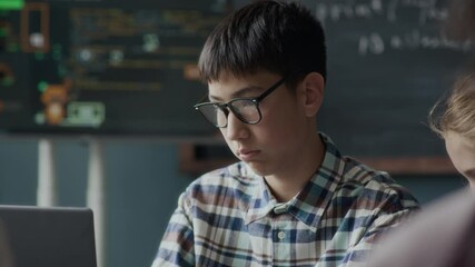 Waist up of Asian teenage schoolboy in glasses and played shirt typing on laptop keyboard while studying coding and programming in class during lesson