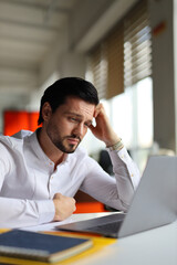 Wall Mural - A man is sitting at a desk with a laptop and a notebook. He is looking at the laptop with a frown on his face