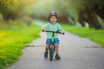 Happy smiling little boy with helmet running and riding on first bike without pedals on sidewalk at city park in warm summer day. Cute 2 years old toddler. Front view. Learning to keep balance.