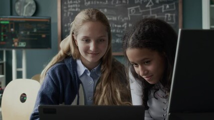 Waist up of two multiracial teenage schoolgirls sitting in front of laptop in modern programming class and having conversation