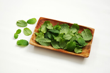 A wooden bowl contains fresh mint leaves, scattered on a white background, with some leaves falling from the bowl.