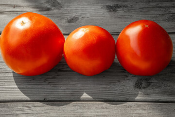 Close-up of three ripe red tomatoes arranged in a row on a rustic wooden surface illuminated by natural light. The wood texture contrasts with the smooth tomatoes.