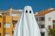 © neiros - In white ghost costume and looking at camera, an unrecognizable kid has fun on the street during the Halloween holiday