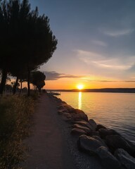 Poster - A serene lakeside path with tall trees and rocks leading to a golden sunset, casting a peaceful reflection on the water at dusk.