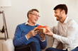 © Prostock-studio - Mature Son Congratulating Elderly Father Giving Him Birthday Gift Sitting On Sofa At Home. Selective Focus