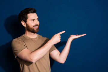 Poster - Photo of cheerful man with beard dressed beige t-shirt look directing at product on arm empty space isolated on dark blue color background