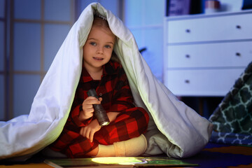 Cute little girl with flashlight reading book under blanket in dark room