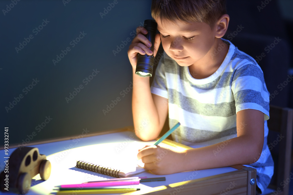 Cute little boy with shining flashlight drawing at table in dark room