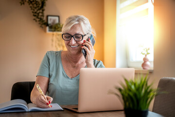 beautiful smiling older senior business woman communicating on mobile phone working at home. one female person work in living room using laptop technologies.