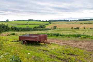 Old farm agricultural trailer with brown painted wood side boards in farmland field and hedgerows