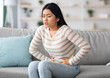 © Prostock-studio - Stomach Ache. Sick Asian Woman Suffering From Acute Abdominal Pain At Home, Upset Korean Female Having Menstrual Pain Or Problems With Digestion, Sitting On Couch And Touching Belly, Free Space