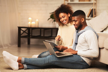 Happy african-american spouses browsing internet on gadgets, using laptop anf tablet at home, copy space