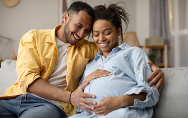 Wall Mural - Pregnancy. Black Husband And Pregnant Wife Hugging Sitting At Home. Family Couple Expecting Baby, Man Embracing Woman Smiling Posing Together. Childbirth Concept