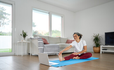 Fit sporty middle aged woman stretching at home looking at laptop. Healthy mature older lady doing sport fitness exercises watching workout body care tutorial in living room of modern house.