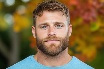 Close-up portrait of a bearded man wearing a blue shirt, standing outdoors with colorful autumn foliage in the background