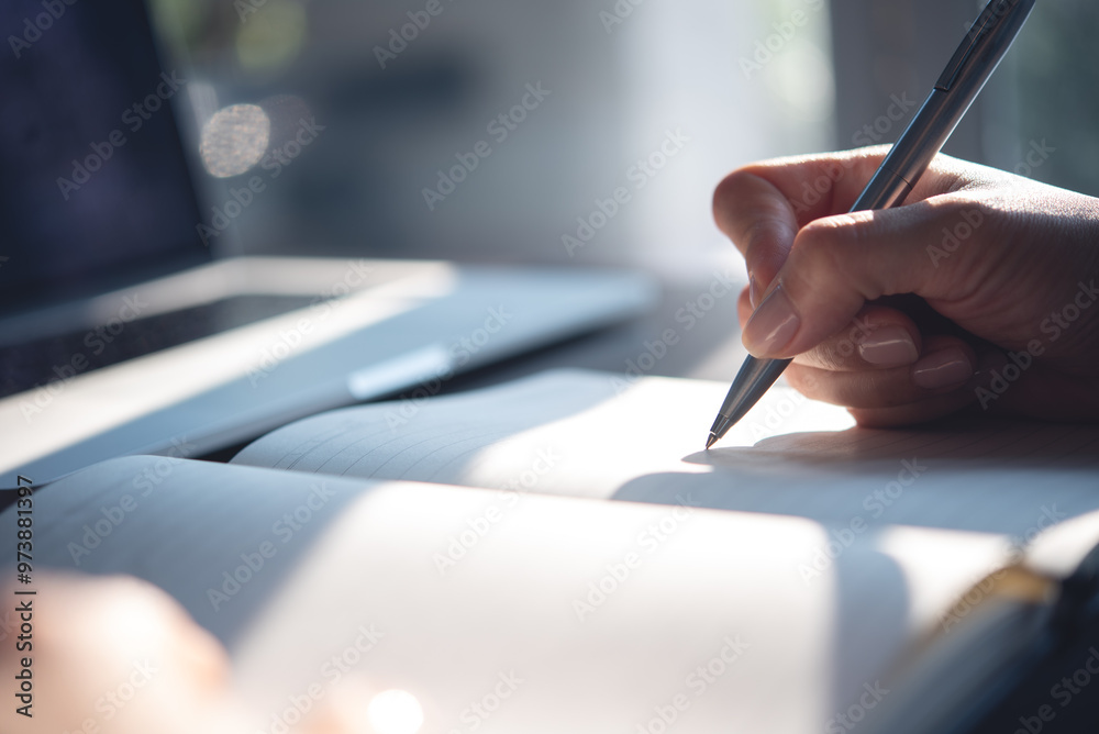 Close up of woman hand with a pen writing on notebook with laptop computer on office table. Business woman planning, making a note reminder. Student studying online, e-learning