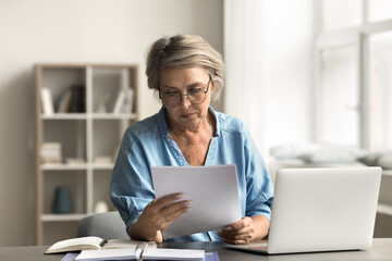 older woman with serious expression read document sitting at desk with laptop, dealing with importan