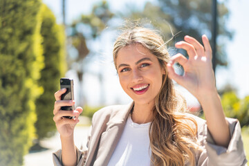 Wall Mural - Pretty blonde Uruguayan woman using mobile phone at outdoors showing ok sign with fingers