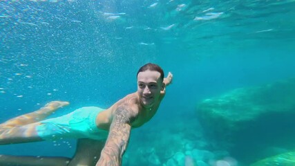 Wall Mural - Close up of one young attractive caucasian man holding camera with hands swimming underwater and enjoying free time on holiday at the beach in the blue sea
