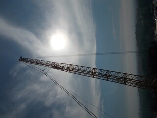 A tall, red communication tower stands against a backdrop of a bright sun and a cloudy sky. The tower stretches high into the air, with cables extending from its peak.