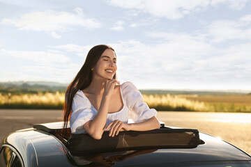 Wall Mural - Enjoying trip. Smiling woman leaning out of car roof outdoors