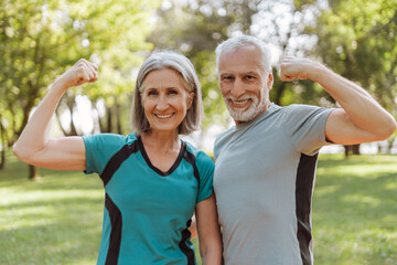 Smiling, attractive senior athletic couple flexing muscles in park, looking at camera outdoors