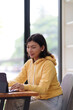 © snowing12 - A young woman in a yellow hoodie is sitting at a table, typing on her laptop. She is working in a bright room with large windows, surrounded by natural light and greenery outside