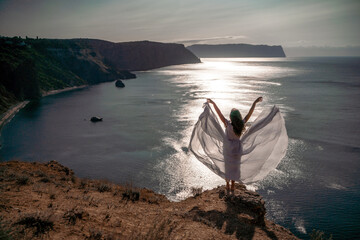 Wall Mural - A woman stands on a rocky cliff overlooking the ocean. She is wearing a white dress and her arms are spread wide, as if she is embracing the beauty of the scene. Concept of freedom and serenity.