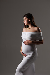 
Beautiful young pregnant brunette girl. A woman waiting for a baby in a white dress with bare shoulders in the studio. Artistic smudges in the photo