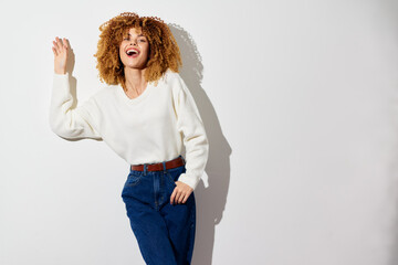 Wall Mural - Happy young woman with curly hair in a white sweater and blue jeans standing against a plain light background, radiating joy and confidence