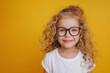 © Victor Bertrand - Smiling child with curly blonde hair, black glasses, and a white top against a vibrant yellow background, showcasing happiness and innocence.