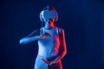 Wall Mural - Female standing surrounded by neon light wear VR headset connecting metaverse, futuristic cyberspace community technology. Elegant woman using both hand interacting with virtual object. Hallucination.